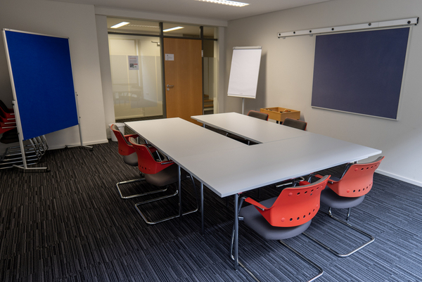 Gruppenraum mit großem weißem Tischequadrat umstellt mit roten Stühlen. An der Wand eine Tafel sowie an der Seite ein blaues Flipchart