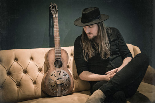 Das Foto zeigt Bad Temper Joe auf einer Couch. Neben ihm eine Gitarre.