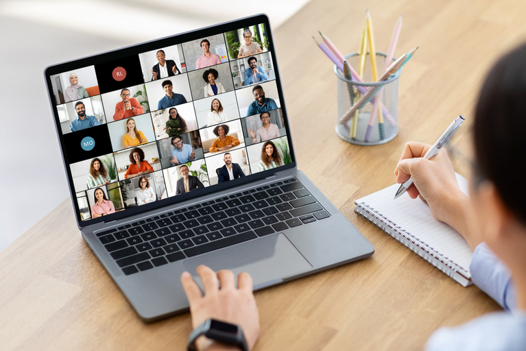 Frau sitzt am Schreibtisch vor Laptop. Auf dem Bildschirm findet eine Online Besprechung mit verschiedenen Teilnehmern statt.