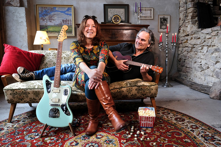 Alexandra Schwarz und Chris Toff Brill auf einem Plüschsofa sitzend in einem retro gestalteten Raum mit Klavier im Hintergrund und Gitarre im Vordergrund